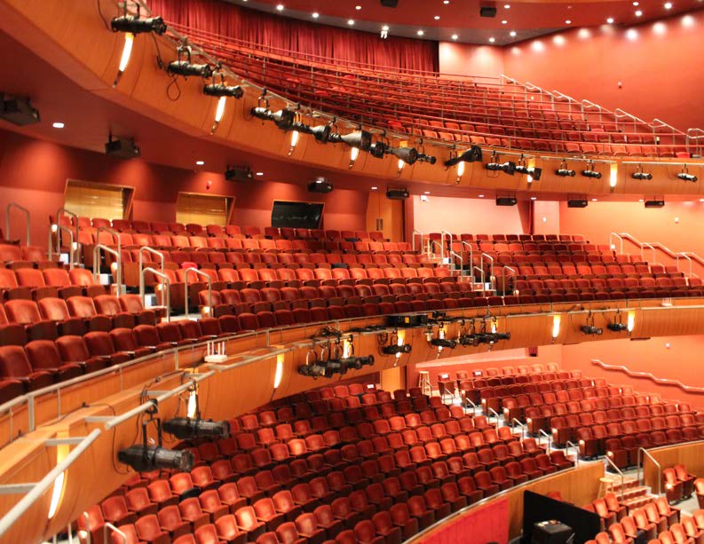 Empty theater auditorium with rows of red seats and balcony lighting rigs.